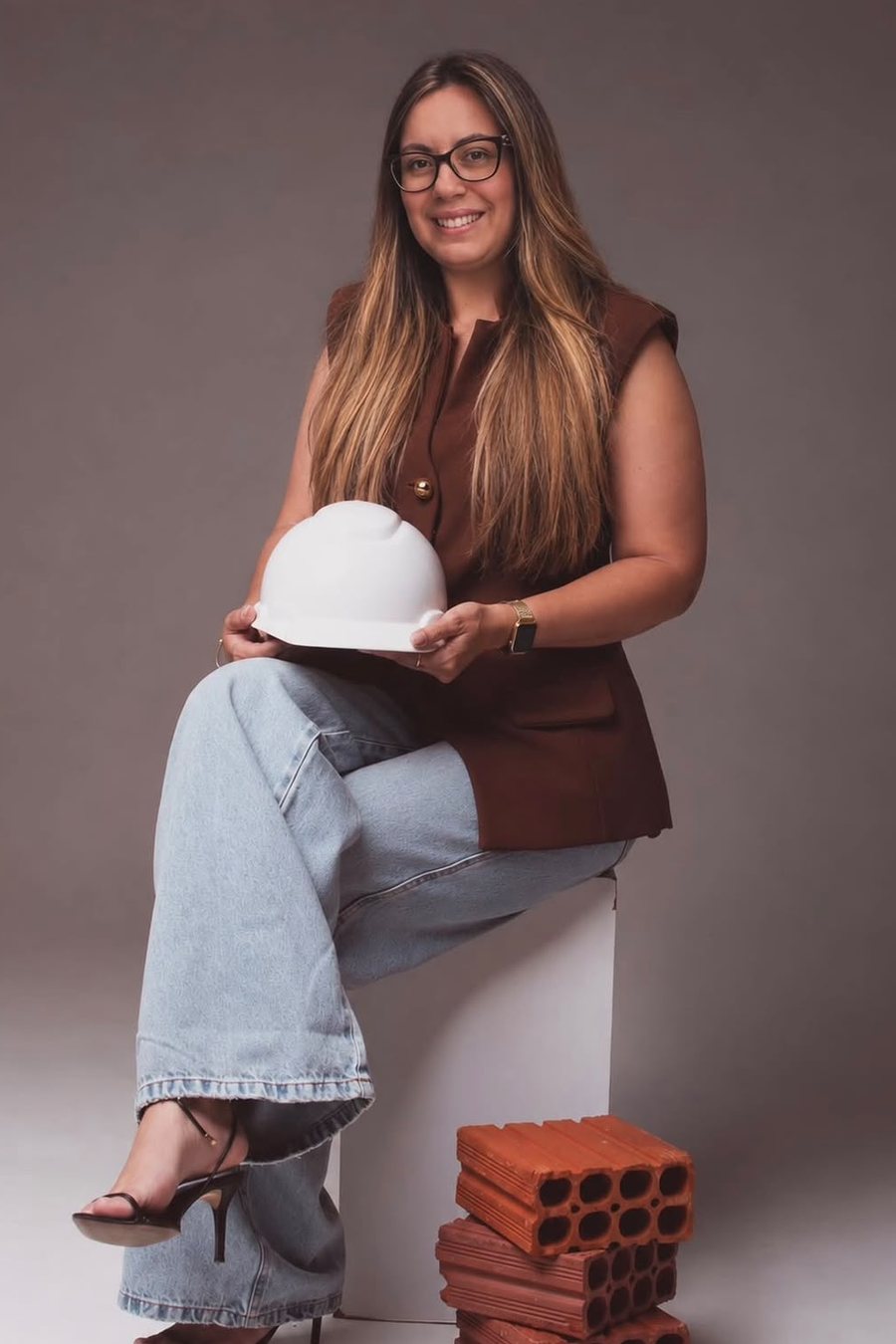 Ana Carolina Garcia sentada em estúdio, segurando um capacete branco de obra, com tijolos cerâmicos ao lado