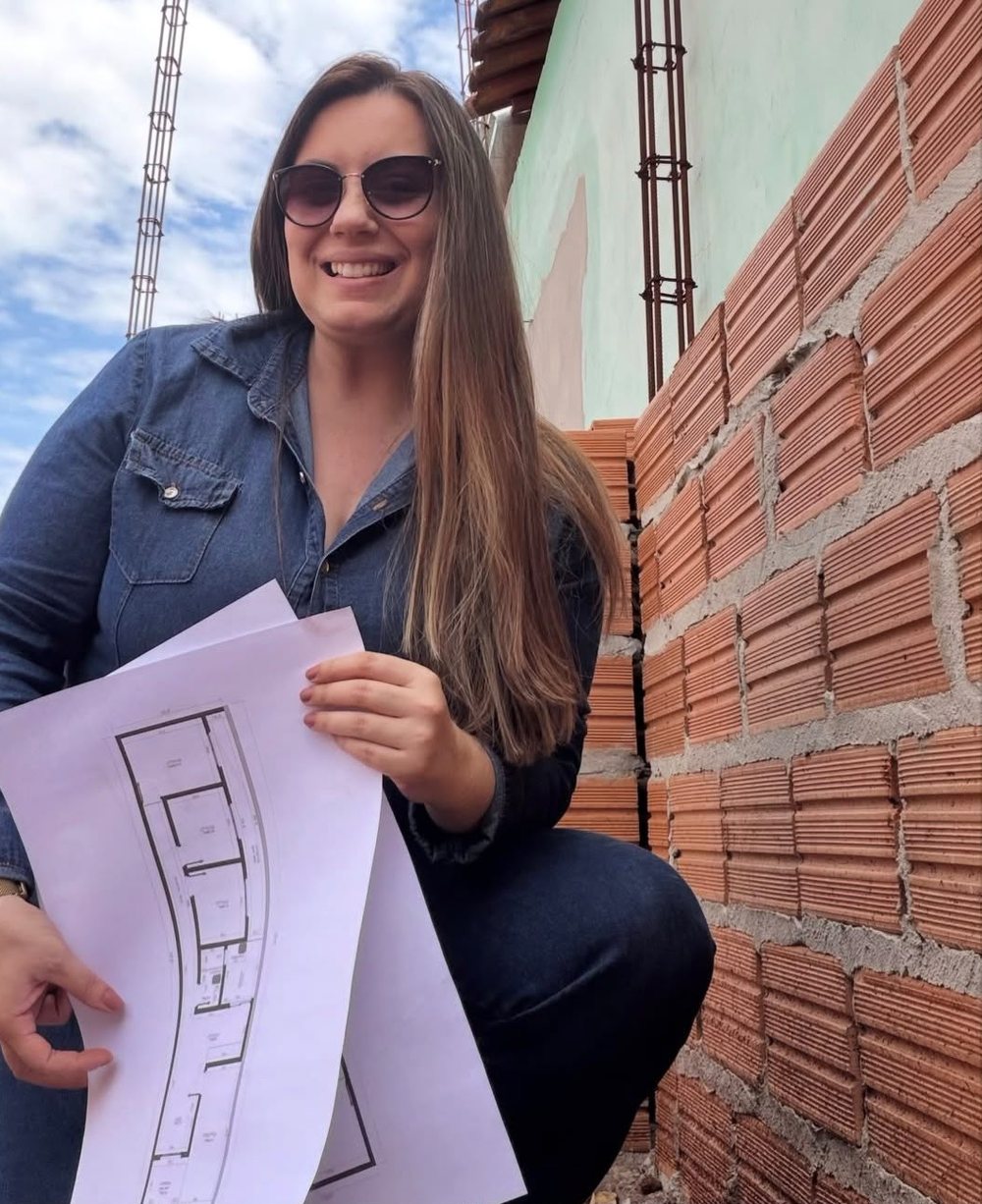 Ana Carolina Garcia em canteiro de obras, agachada ao lado de uma parede de tijolos, mostrando a planta baixa impressa